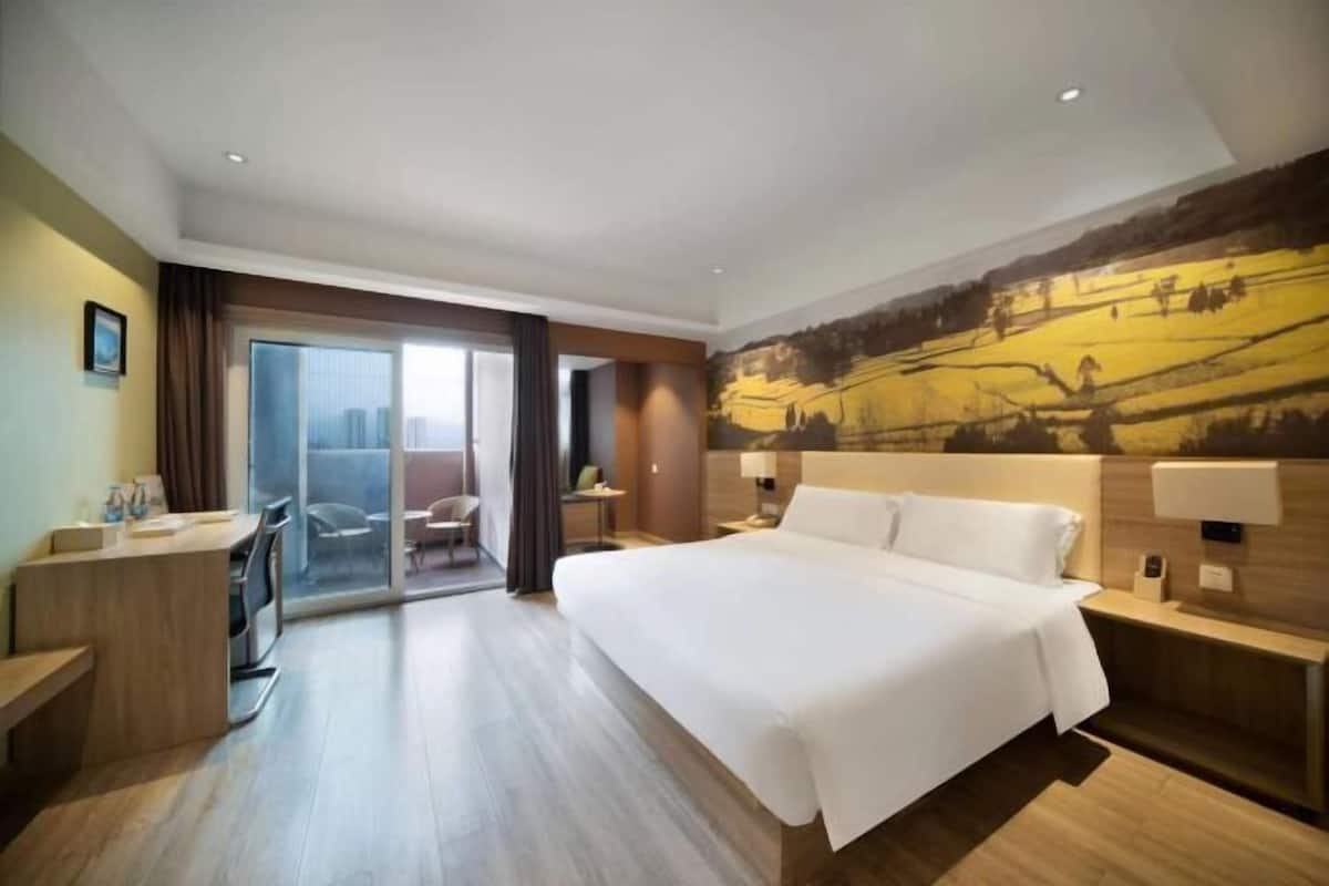 A hotel room with a large bed, a desk, a chair, a small table, and a view of a cityscape through a glass door.