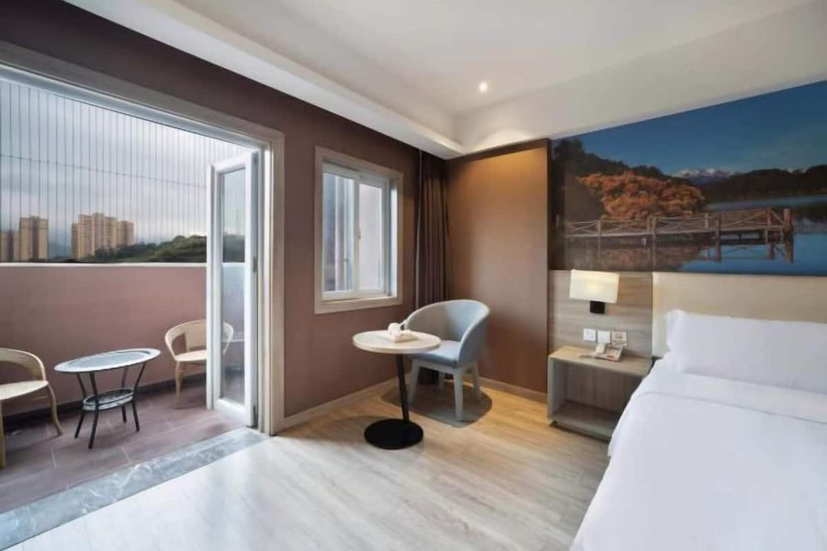 A hotel room with a bed, a desk, a chair, a small table, and a painting of a lakeside scene.