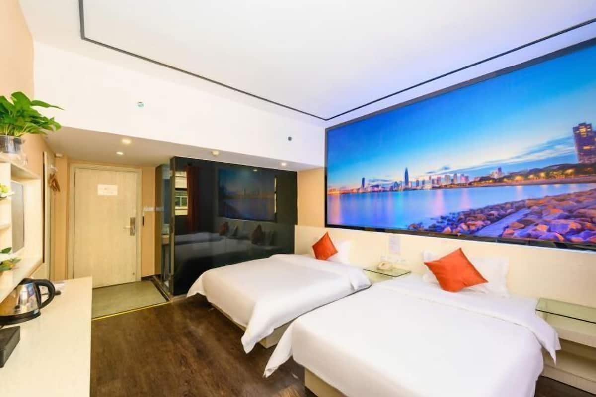 A hotel room with two beds, a large cityscape painting on the wall, a desk with a kettle, and a potted plant.