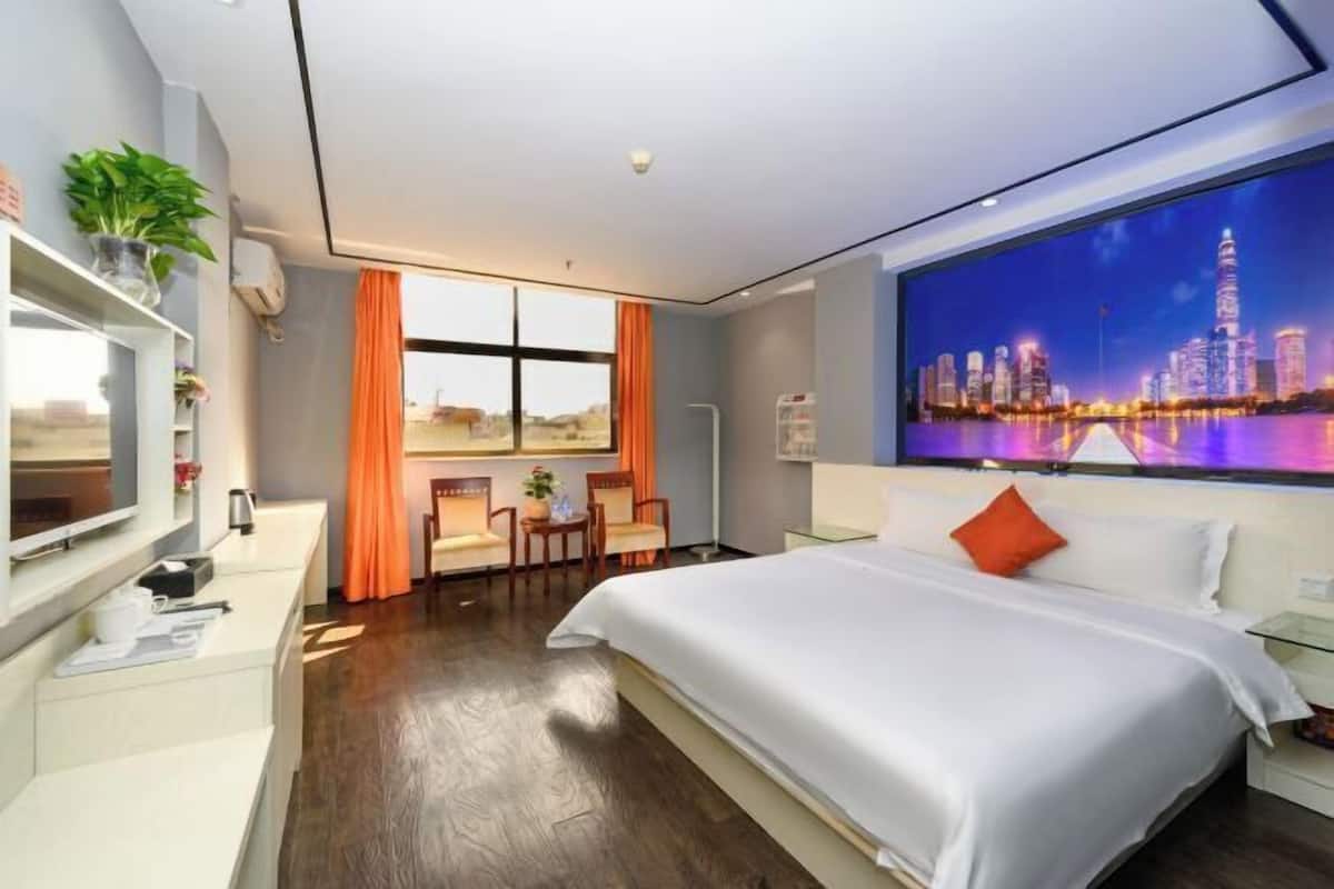 A modern hotel room with a large bed, a desk with a chair, a large window with curtains, and a cityscape view on the wall.