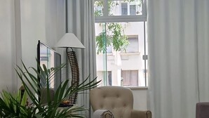 A modern living room with a beige armchair, a black rug, a potted plant, and a large window with curtains.