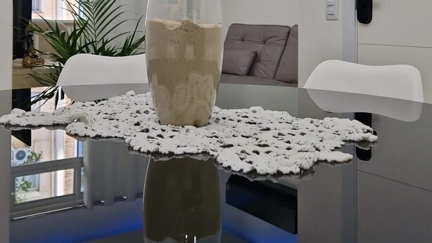 A modern living room with a glass-top dining table, a clear vase with sand, a lace table runner, a grey sofa, and a white door.