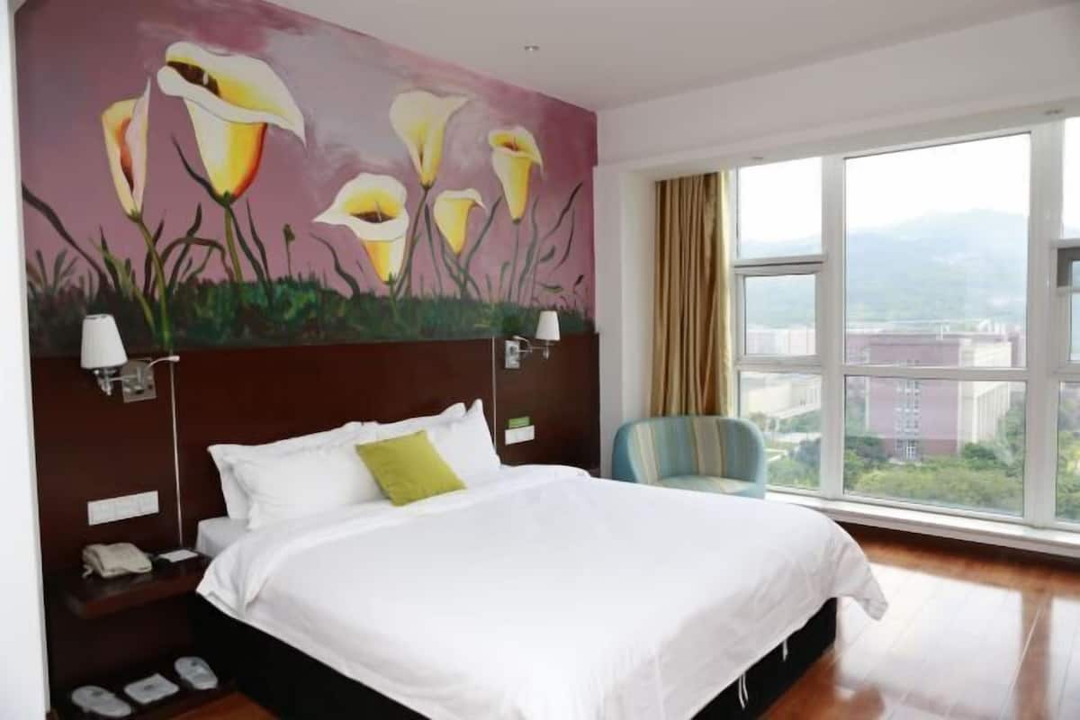 A hotel room with a large bed, a nightstand, a lamp, a chair, and a window with a view of buildings and greenery.