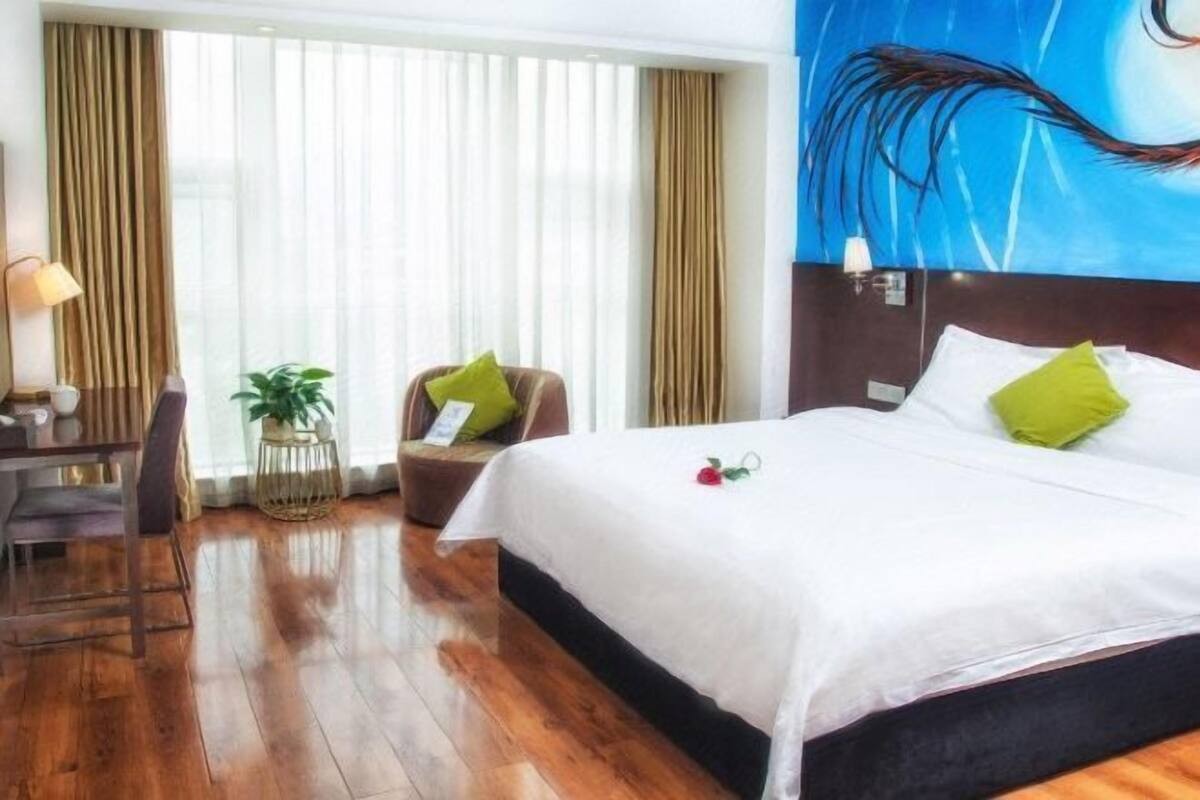 A hotel room with a wooden floor, a bed with white bedding and green pillows, a desk with a lamp, and a chair.
