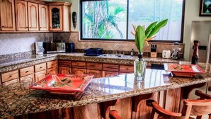 A kitchen with wooden cabinets, a granite countertop, a large window with a view of greenery, and a central island with chairs.