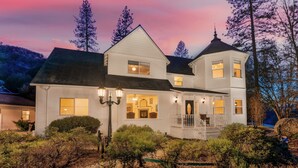 A large, white, two-story house with a dark roof, multiple windows, and a porch with a railing.