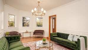 A spacious living room with a green sofa, a brown leather sofa, and a green velvet sofa. There is a glass coffee table with a plant on it. The room has large windows with views of trees and a wooden door.