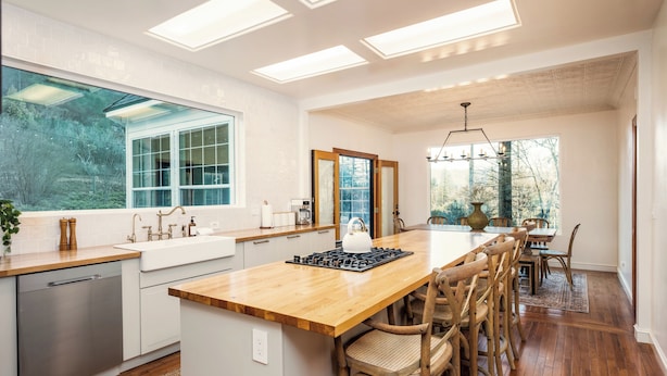 A modern kitchen with a central island, wooden chairs, and a dining area with a view of trees.
