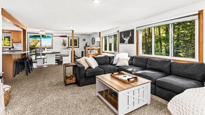 A modern living room with a sectional sofa, a coffee table, and a view of the outdoors through large windows. - Lakefront Cabin, Hot Tub, Sauna, Fireplace, Game Room, Near Duluth  (Saginaw)