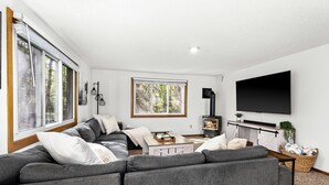 A modern living room with a grey sectional sofa, a flat-screen TV mounted on the wall, and a wood-burning stove. - Lakefront Cabin, Hot Tub, Sauna, Fireplace, Game Room, Near Duluth  (Saginaw)