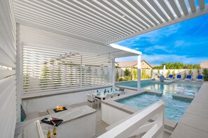 Un área de piscina moderna al aire libre con una zona de estar cubierta, sillas de descanso y una chimenea.