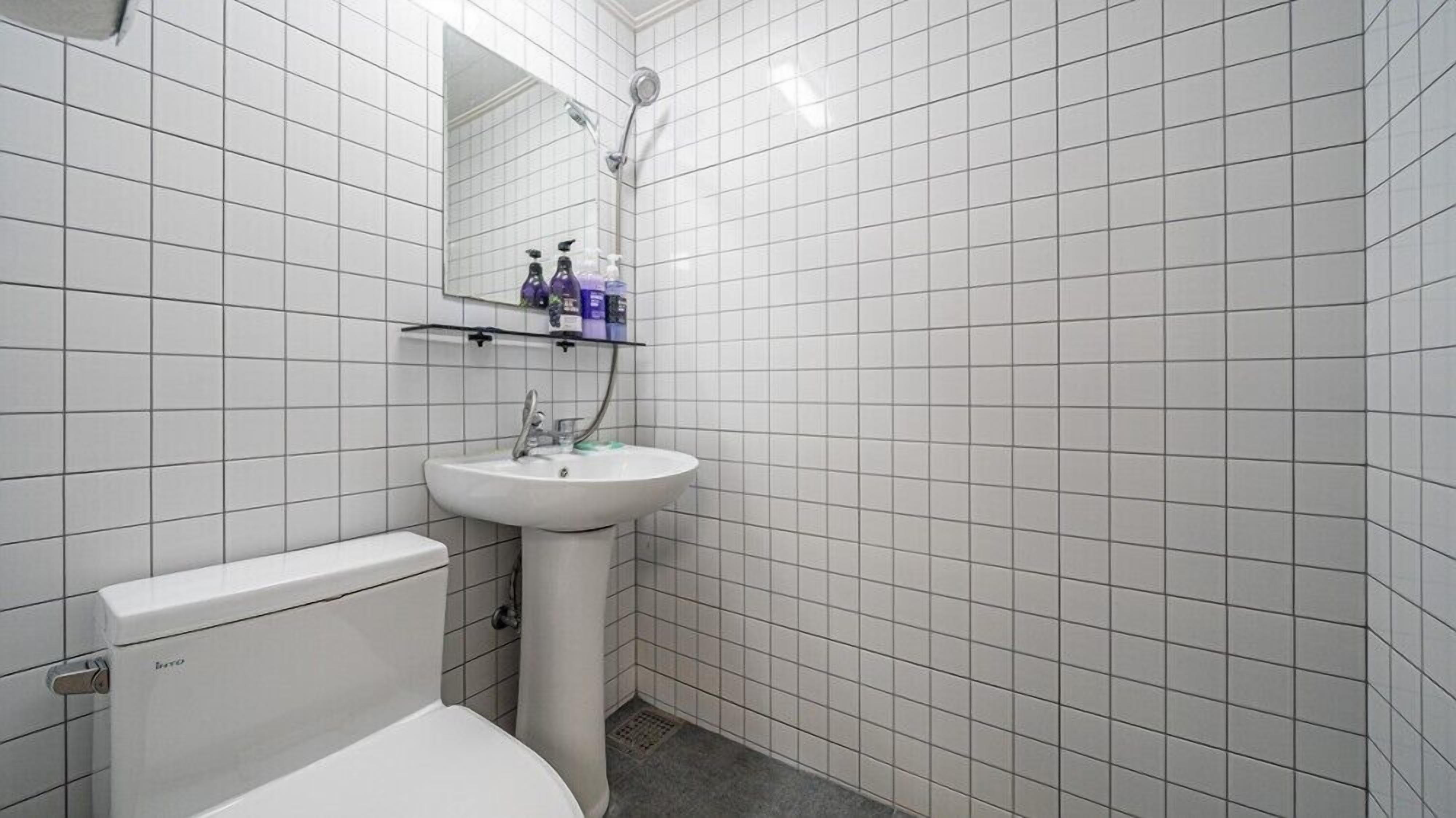 Et badeværelse med vægge af hvide fliser, en hvid vask, et toilet og en brusekabine. Der er hylder med toiletartikler over vasken.