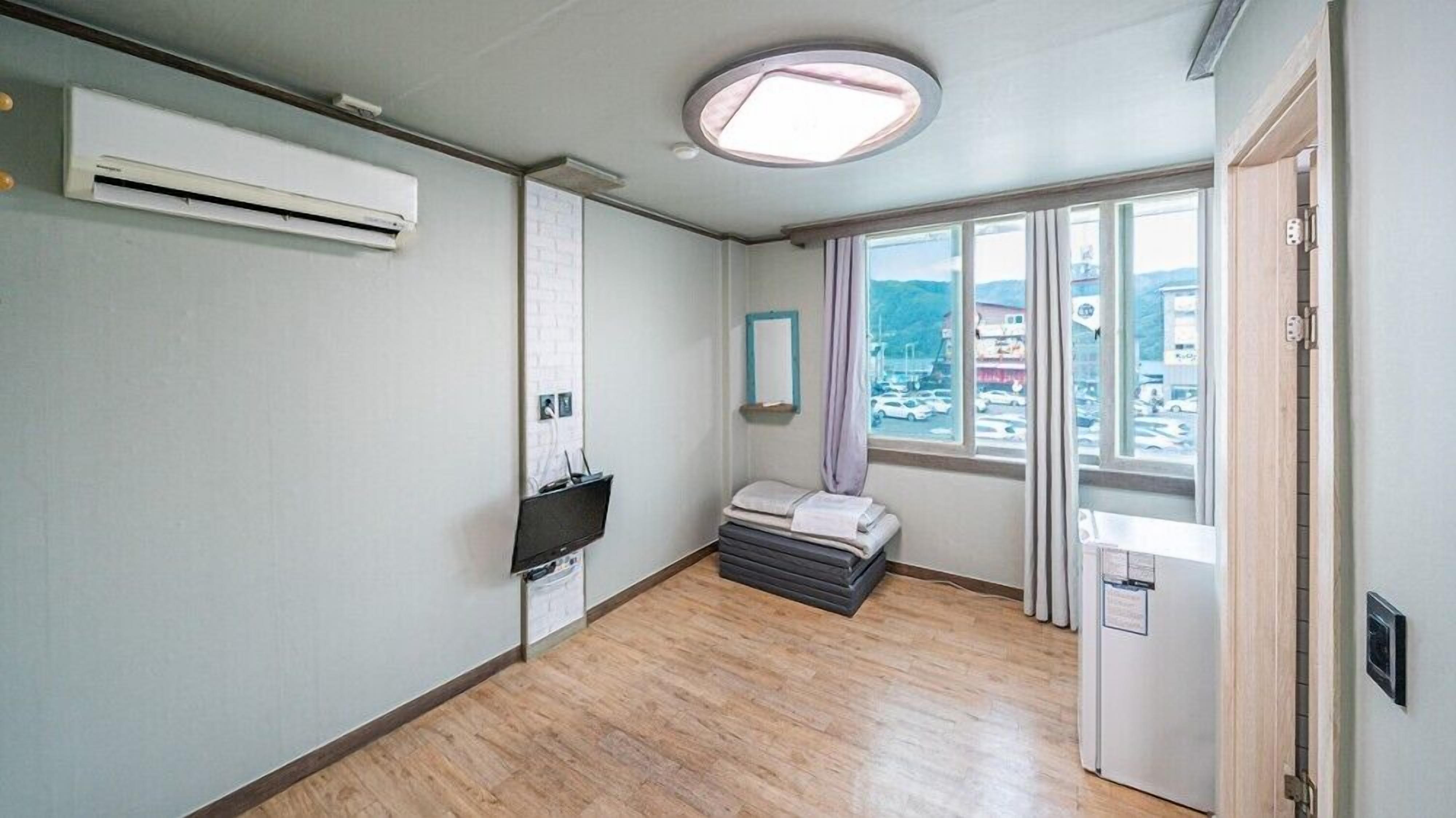 A hotel room with a bed, a small table, a window with a view of the city, an air conditioning unit, and a door.