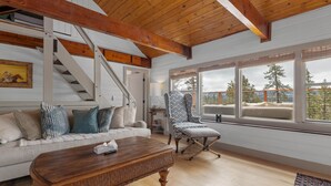 Un salón amplio con techo de madera, una escalera, un ventanal con vistas a árboles, un sillón estampado y un sofá blanco con cojines.