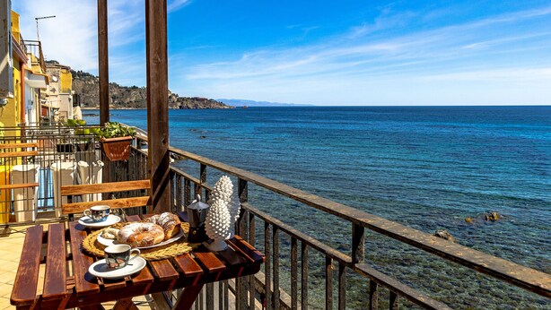 A wooden deck with a table set for breakfast, overlooking a clear blue sea and a coastal town. - Aquamarina Home Seafront House Giardini Naxos (Naxos)