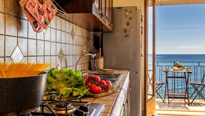 A kitchen with a stove, pots, and fresh vegetables. There is a view of the sea through an open door. - Aquamarina Home Seafront House Giardini Naxos (Naxos)