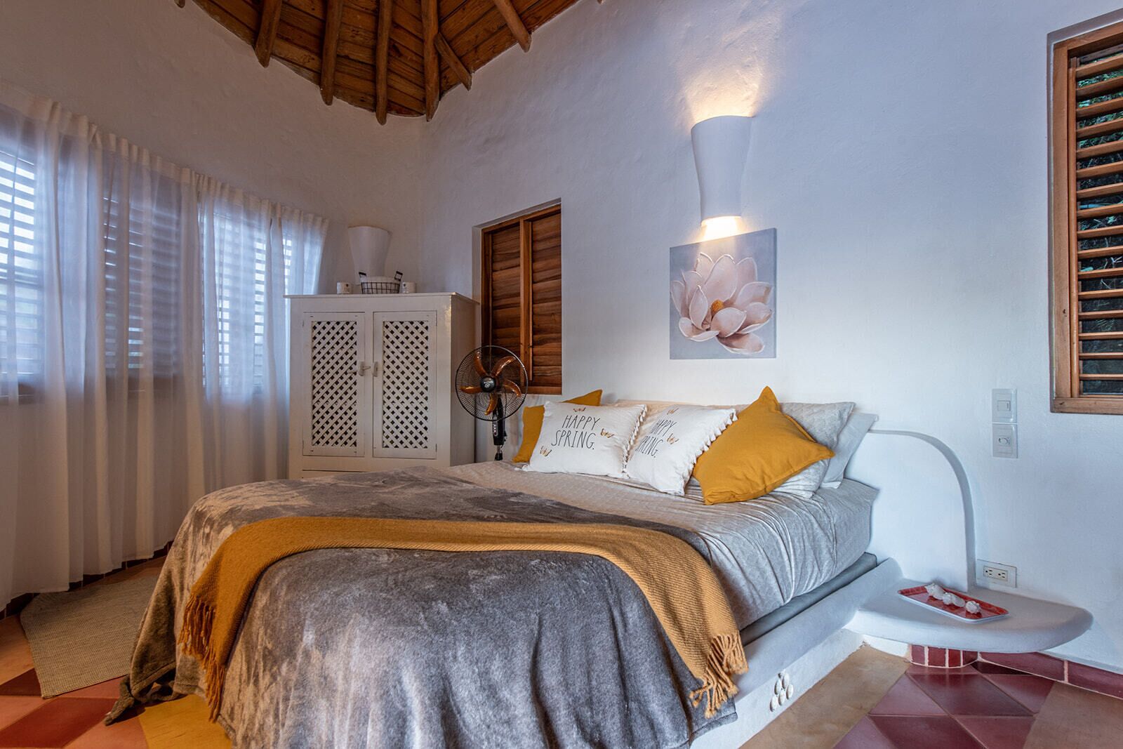 Un dormitorio con cama, mesita de noche, lámpara, ventilador, armario y ventana con cortinas.