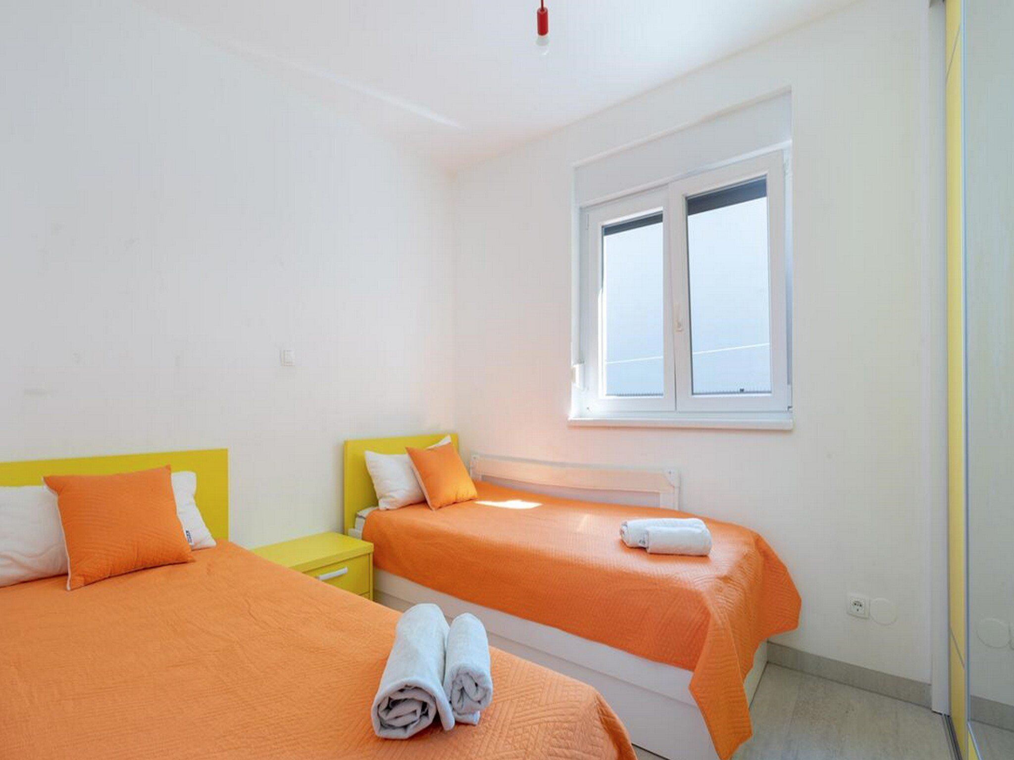 A hotel room with two beds, each with an orange bedspread and white pillows. There is a window with white frames, and a yellow nightstand with a white lamp.