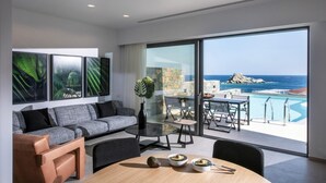 Ein modernes Wohnzimmer mit einem grauen Sofa, einem Glastisch und einem Essbereich mit Holztisch und Stühlen. Ein großes Fenster bietet Blick auf das Meer und eine felsige Landzunge.