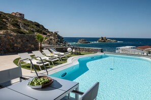 A pool area with lounge chairs, a table with a bowl of apples, and a view of the sea and rocky outcrops. - Made By The Sea Villas 21 Bdrm (Malevizi)