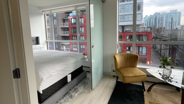 A modern bedroom with a bed, a chair, and a view of a cityscape through large windows. - Trendy Chinatown apartment with parking (Vancouver)