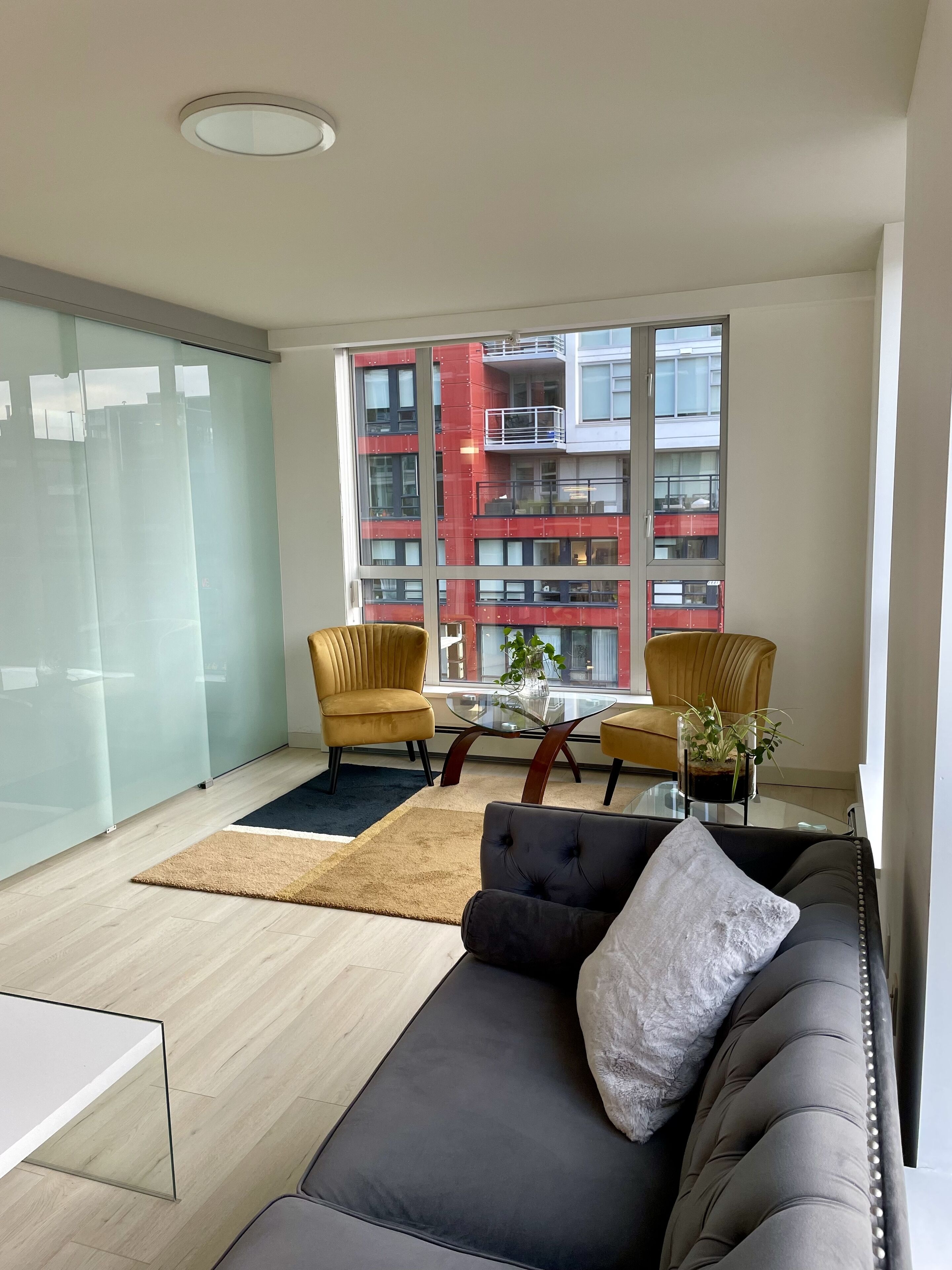 A modern living room with a glass partition, a sofa, two armchairs, a coffee table, and a view of a red building.