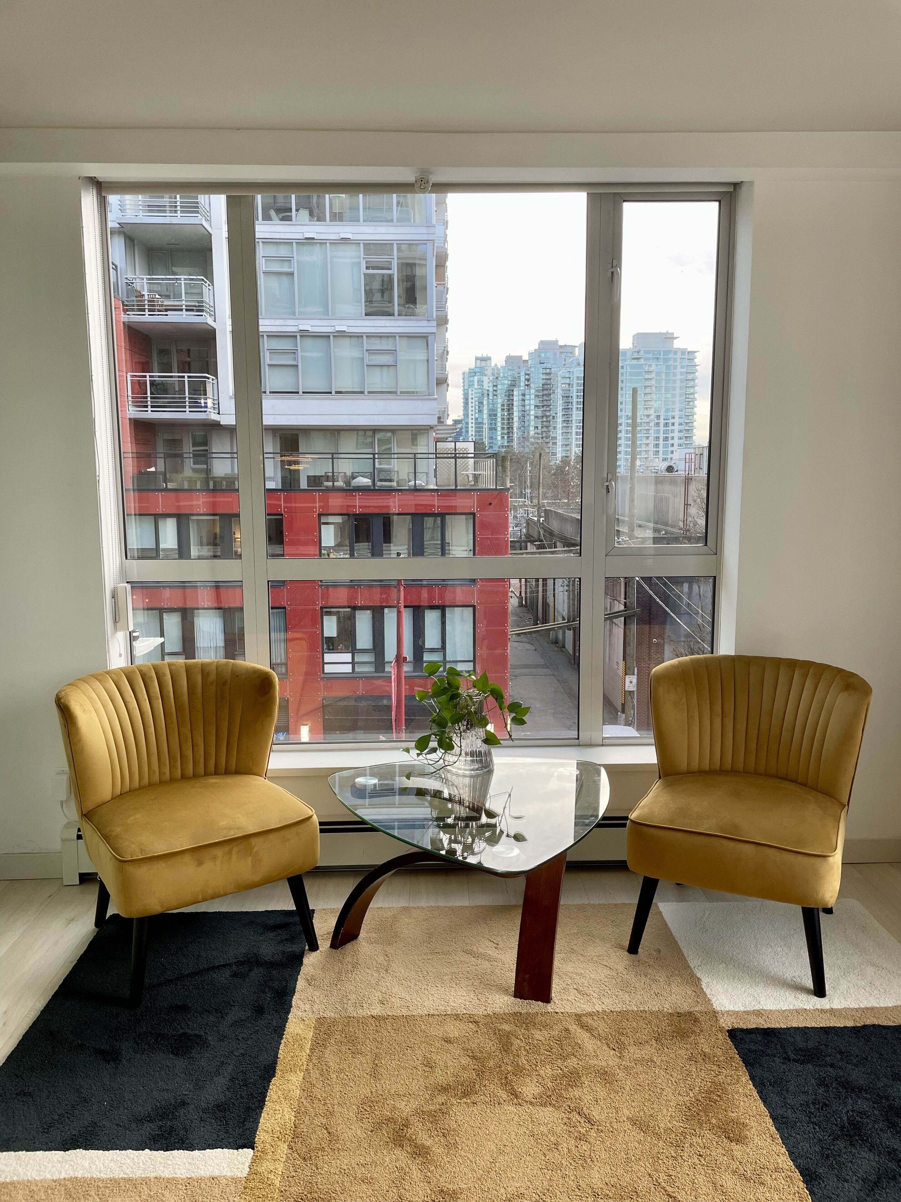 Un salon moderne avec deux fauteuils jaunes, une table basse en verre et une vue sur un immeuble rouge, visible à travers de grandes fenêtres.