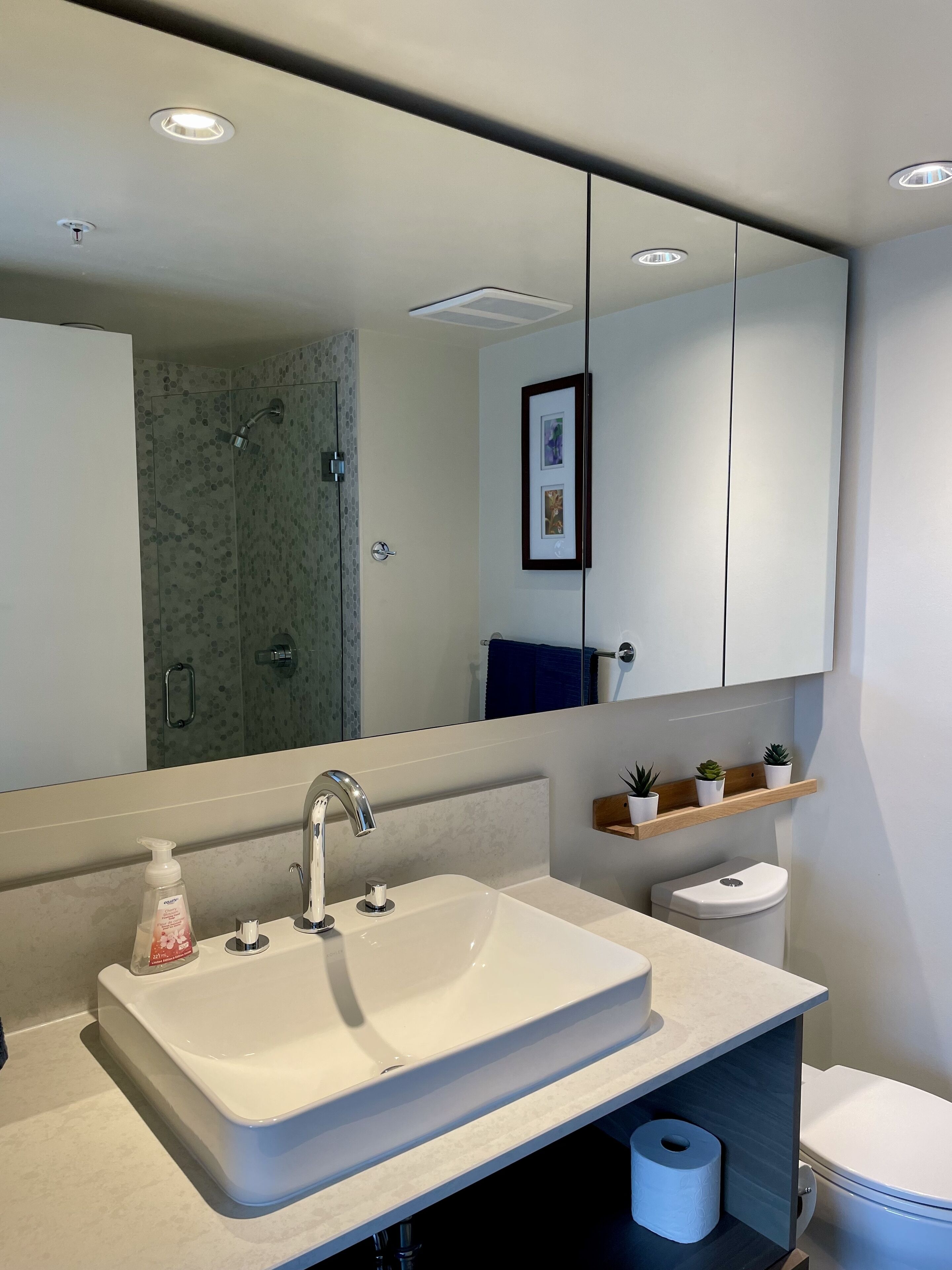 Un baño moderno con un espejo grande, un lavabo con dispensador de jabón, inodoro y zona de ducha.