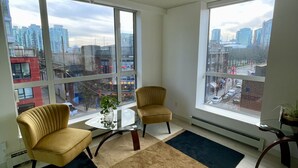 A modern living room with large windows, two armchairs, a small table, and a potted plant. - Trendy Chinatown apartment with parking (Vancouver)