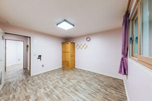 Una habitación amplia con suelo de madera en patrón de espiga, un armario de madera, un pequeño reloj de pared y una ventana con cortinas moradas.