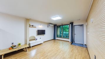 Une pièce spacieuse dotée d’un plancher en bois, d’un téléviseur blanc fixé au mur, d’un banc et d’une porte-fenêtre coulissante donnant sur un espace extérieur.