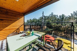 Una terraza de madera con una mesa, sillas y baranda de cristal, con vista a árboles.