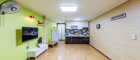 A hallway with a TV, a fan, a small table, and a kitchenette area.