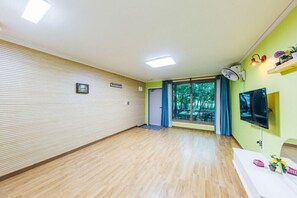 A spacious room with a wooden floor, a large window with blue curtains, a flat-screen TV mounted on the wall, and a small shelf with decorative items. - Jangsa Solbat Pension (Yeongdeok)