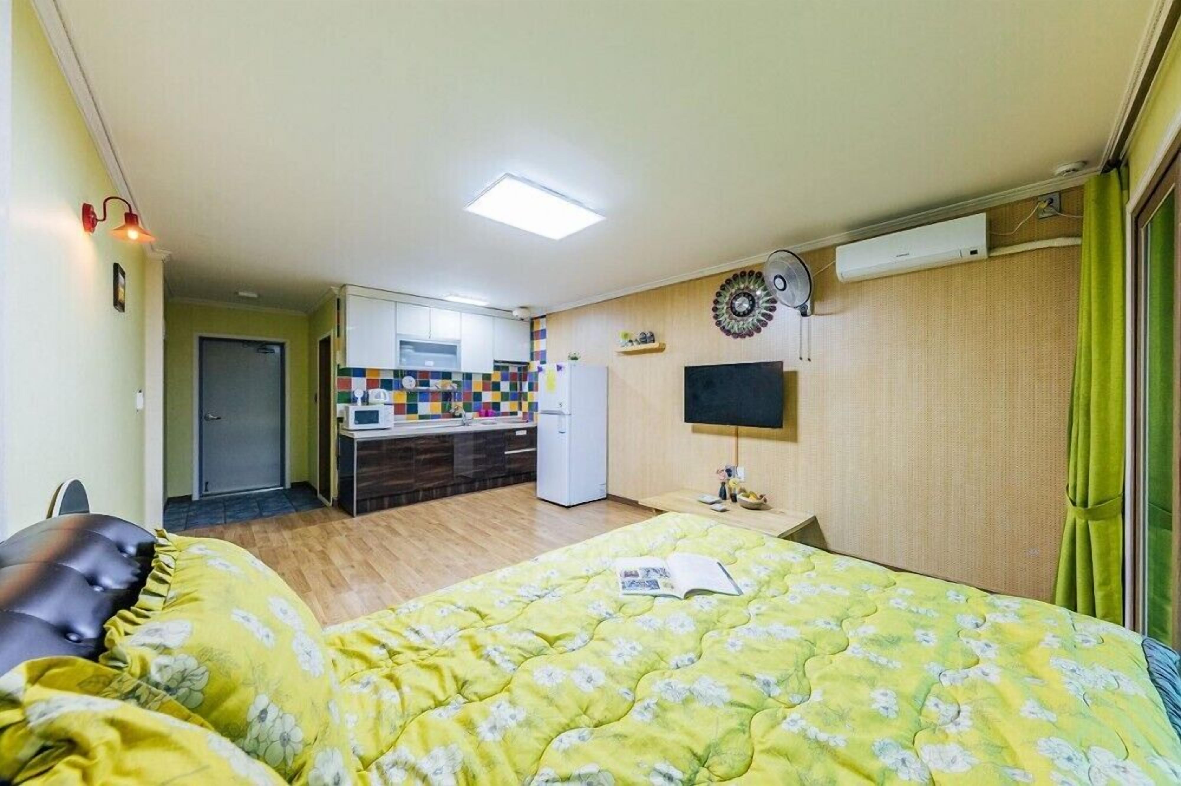 A compact hotel room with a bed, a small desk, a TV, and a kitchenette in the background.