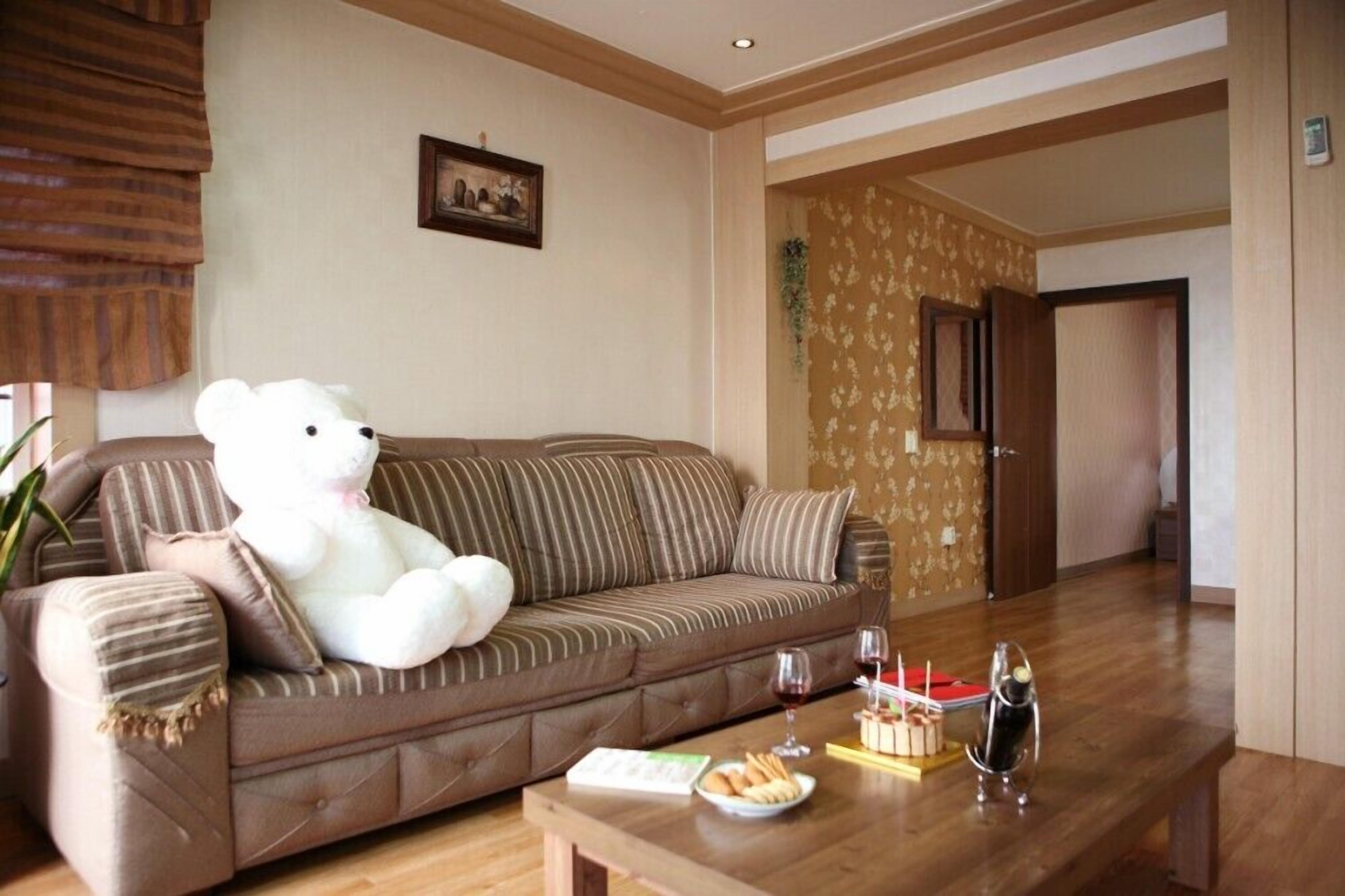 A living room with a large white teddy bear on a brown sofa, a wooden coffee table with snacks and drinks, and a door leading to another room.