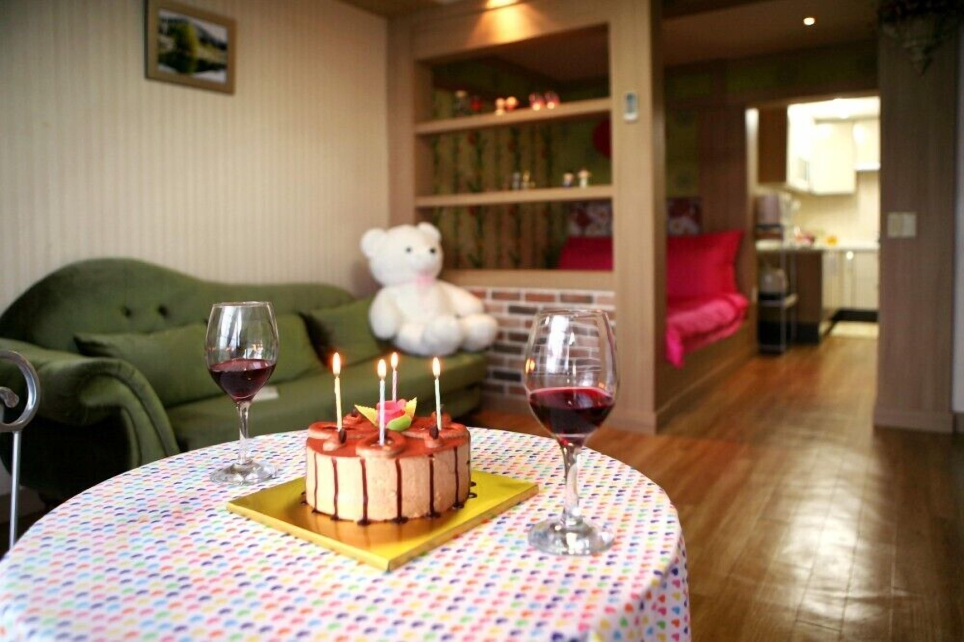 A cozy living room with a green sofa, a teddy bear, a birthday cake with candles, and two glasses of red wine on a round table.