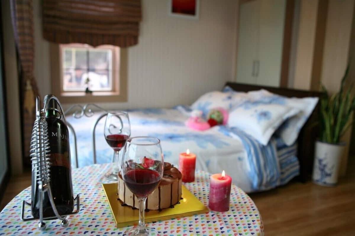 A bedroom with a bed, a bedside table with a bottle, a glass of red wine, and a candle.