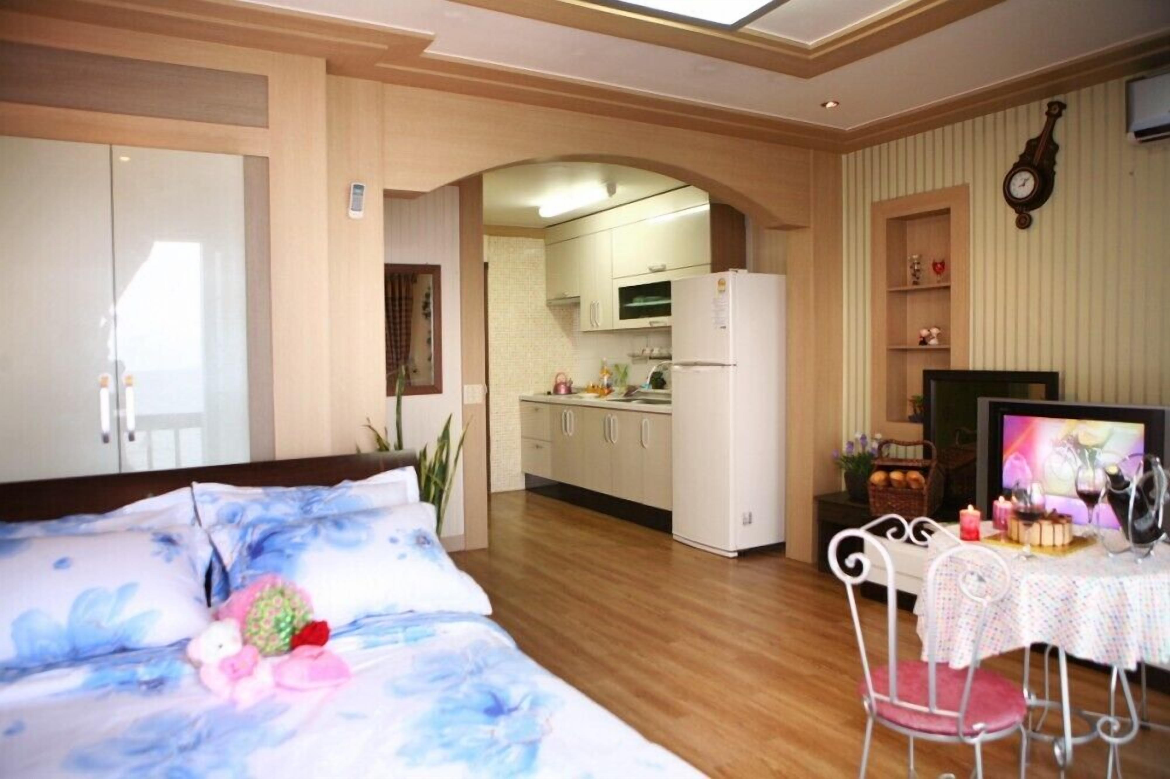 Ein Schlafzimmer mit einem Bett, einem kleinen Tisch mit Fernseher und einer Küchenzeile im Hintergrund.