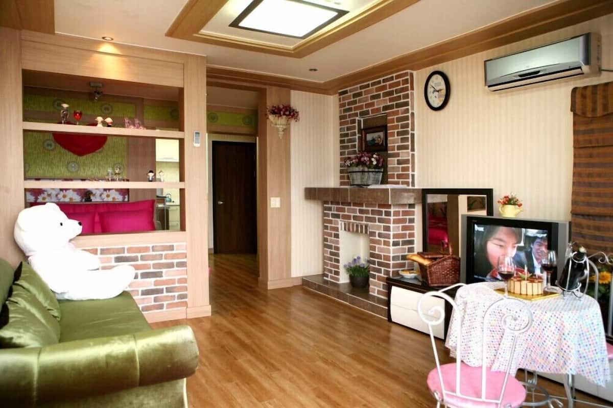 A living room with a green sofa, a white teddy bear, a brick fireplace, a television, and a dining table with a cake.