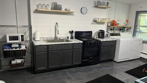 Una cucina moderna con mobili scuri, lavello in acciaio inossidabile, forno a microonde, piano cottura e lavastoviglie. Sopra il bancone, mensole con piatti e bicchieri.