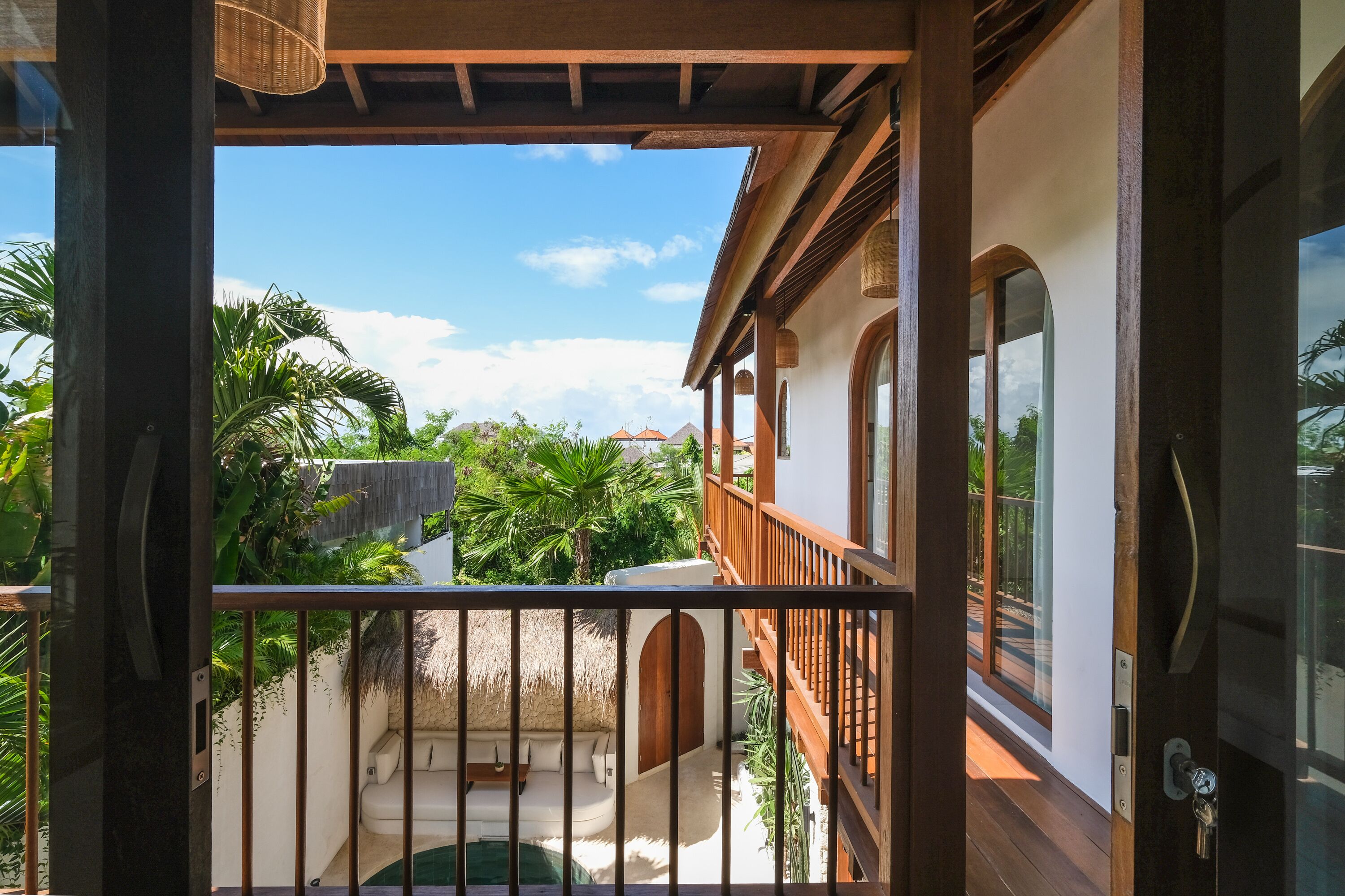 A balcony with a view of a pool and tropical plants, wooden railings, and a thatched-roof structure.