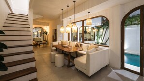 A modern interior with a staircase, a dining area with a wooden table and white sofas, and a view of a swimming pool. - The Arki Villa (Canggu)