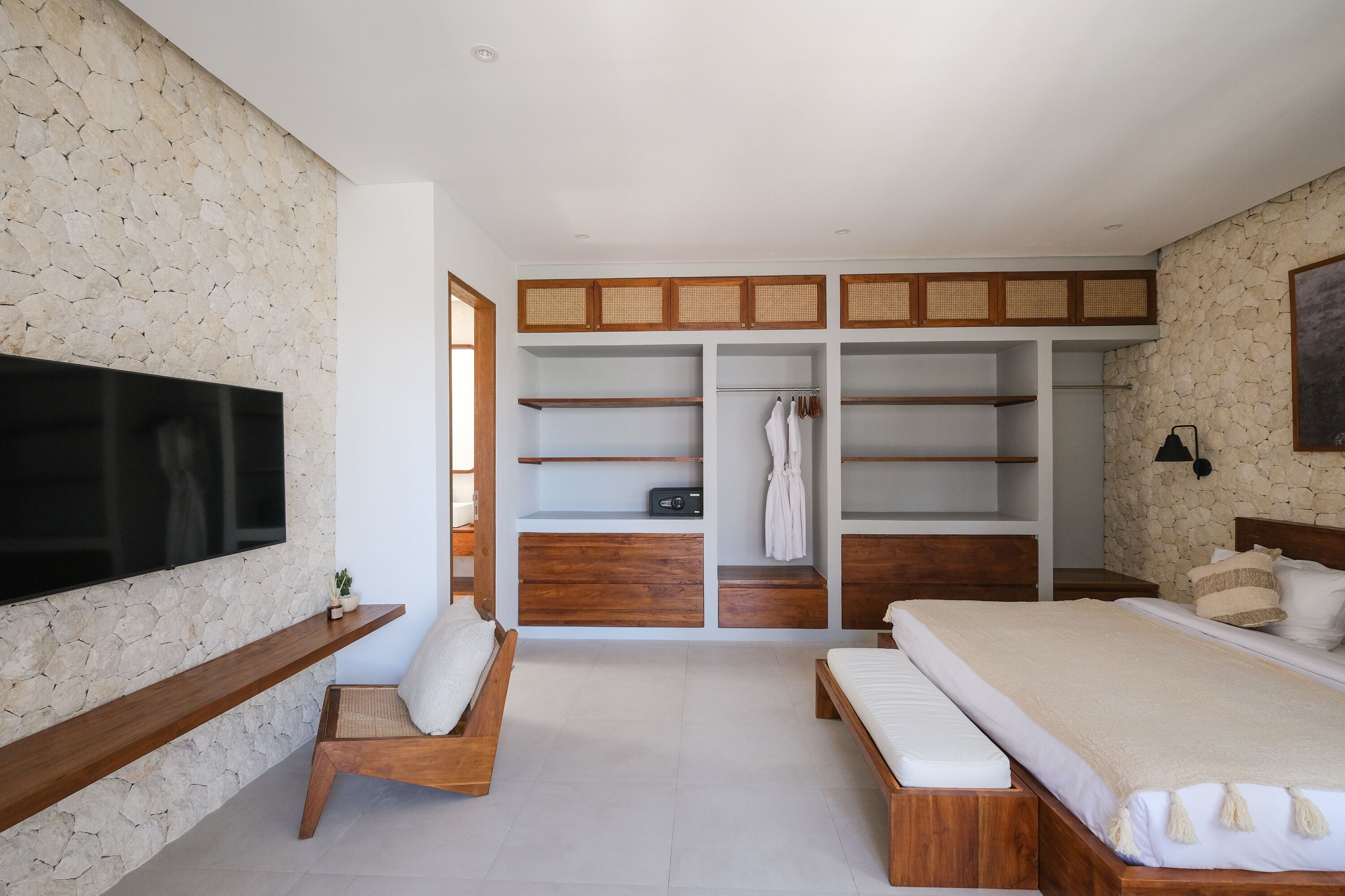 Un dormitorio con una cama grande, un televisor montado en una pared de piedra, un banco de madera y estanterías empotradas.