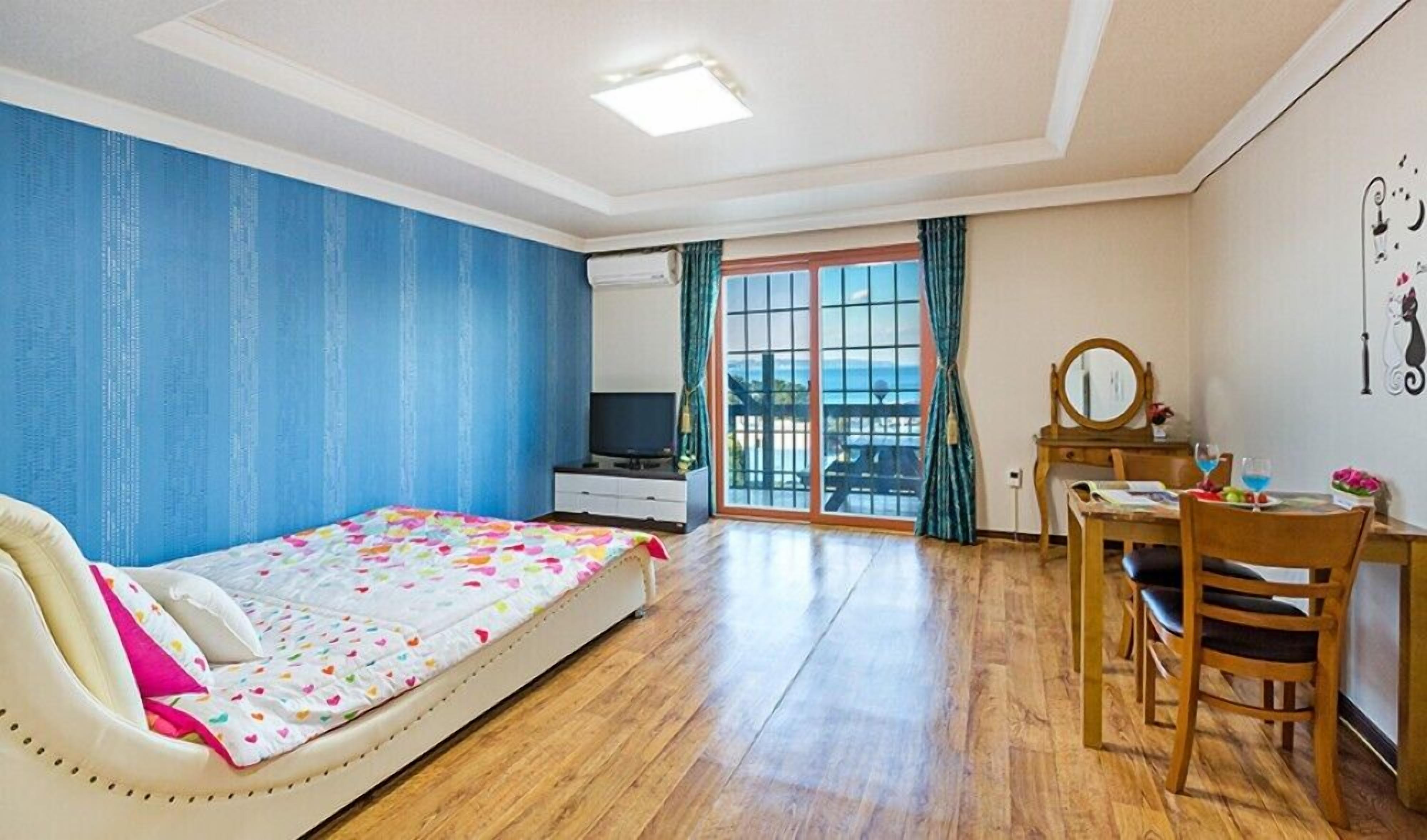 A bedroom with a bed, a desk, a chair, a TV, and a view of the sea through a glass door.