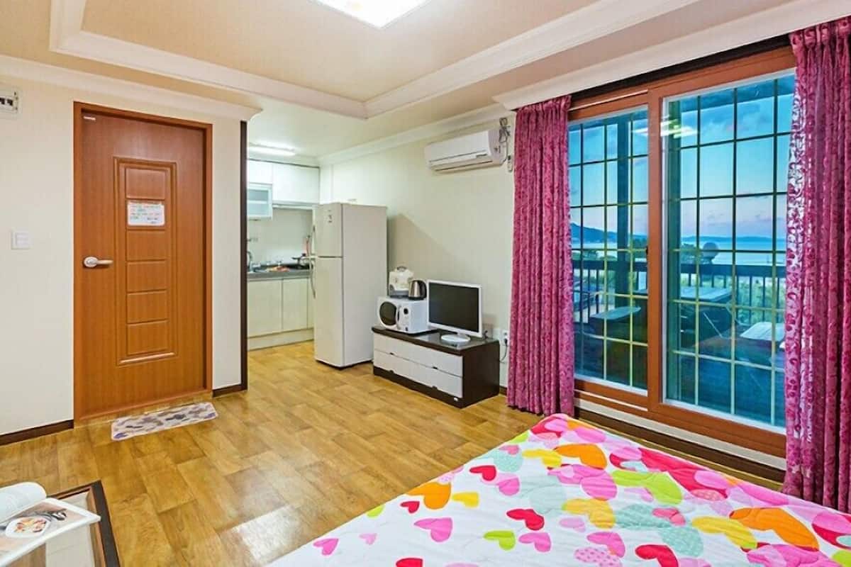 A bedroom with a large window, a bed with a colorful quilt, a small kitchenette, and a wooden door.