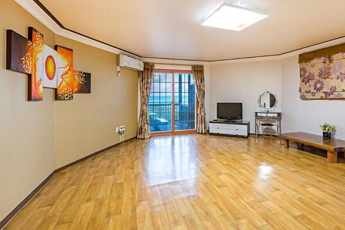 A spacious living room with a wooden floor, a television, a small table, and a large window with curtains.