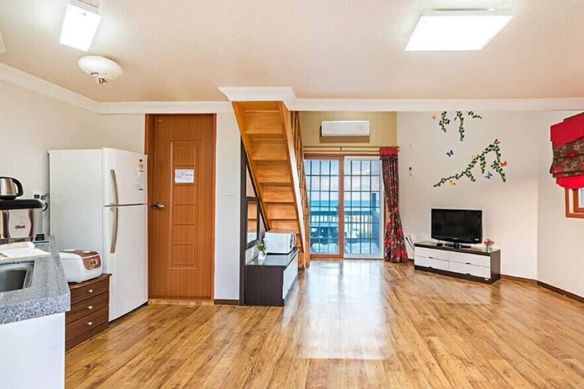 A spacious living area with a wooden floor, a kitchen with a refrigerator, a staircase, and a TV on a stand.