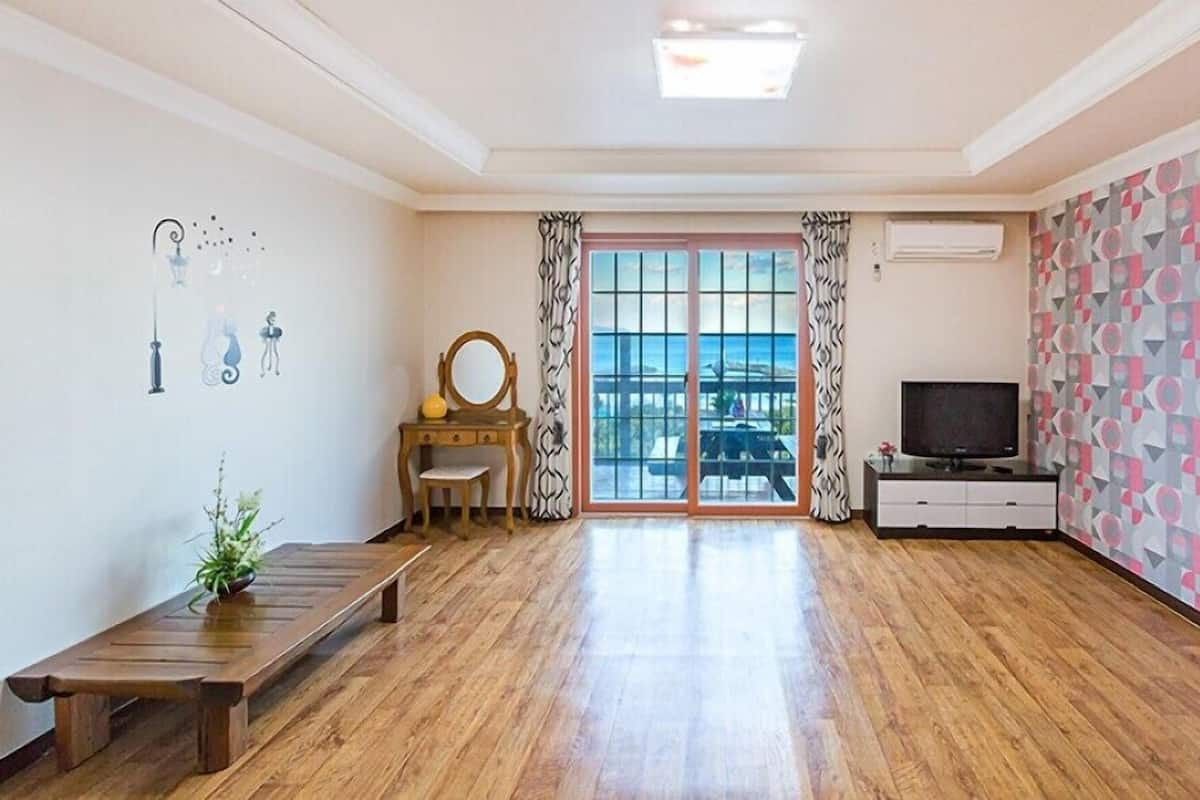 A spacious room with a wooden floor, a wooden bench, a small table with a mirror, a television on a white cabinet, and a large window with curtains.