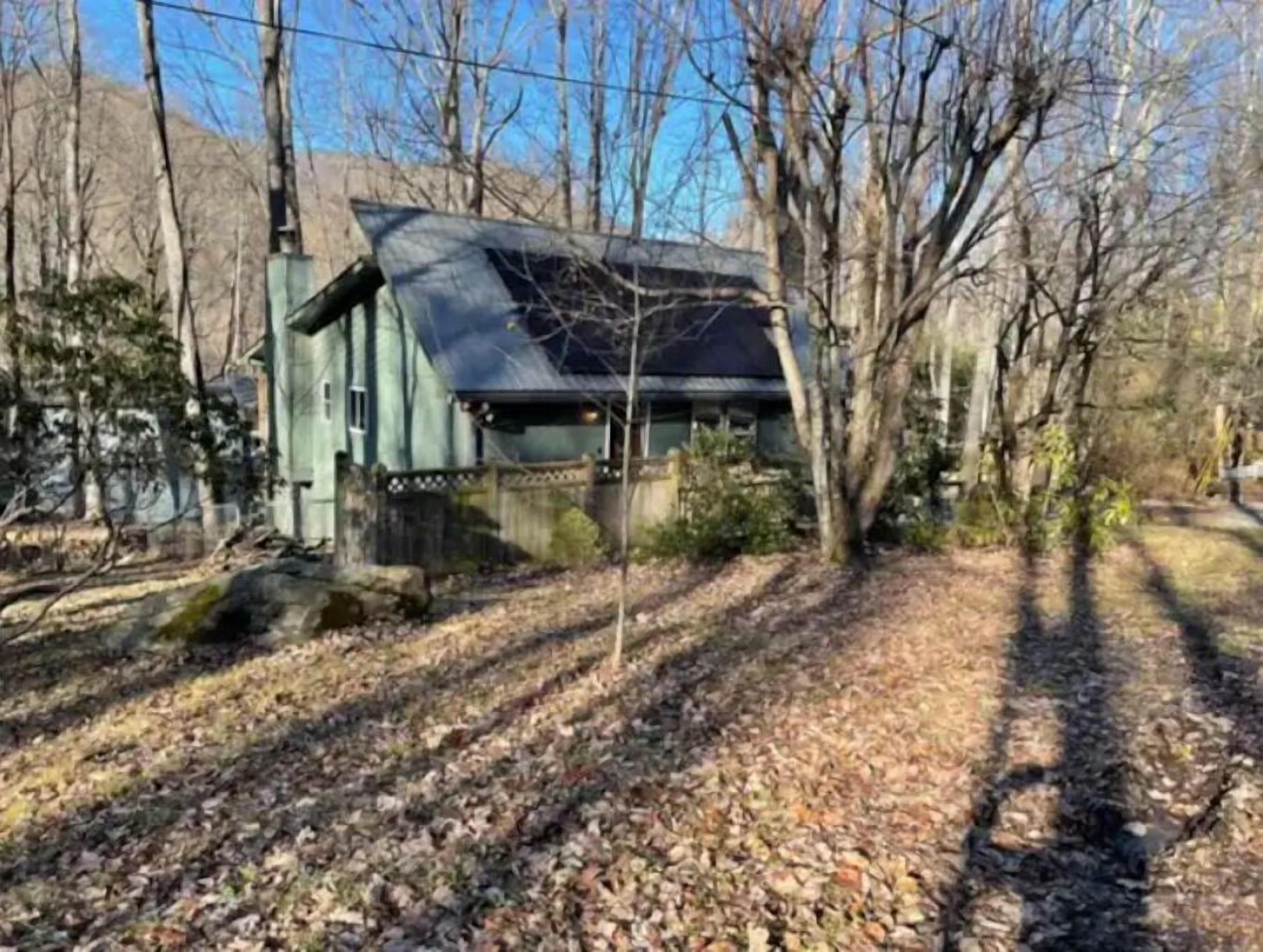 A rustic cabin with a sloped roof, surrounded by leaf-covered ground and bare trees.